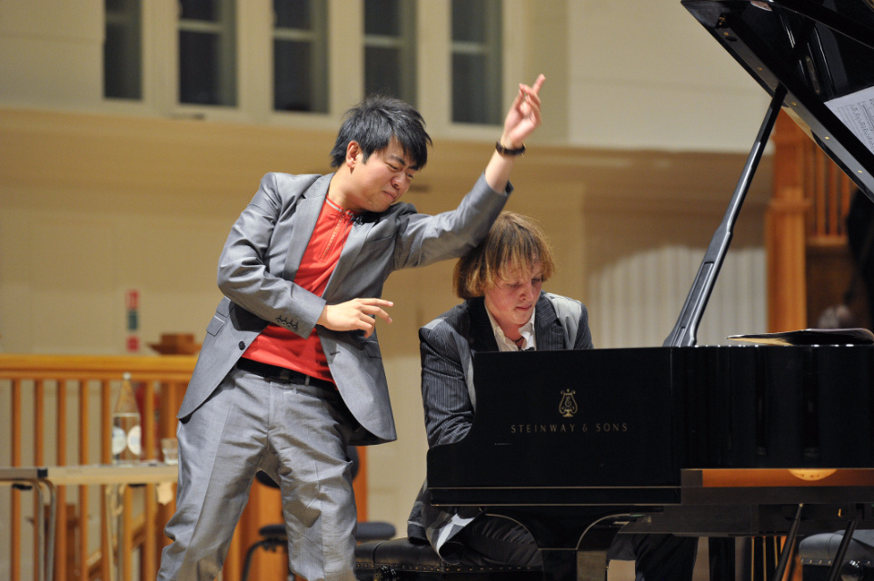 An Asian man teaching a white student, performing on a grand piano, on stage.
